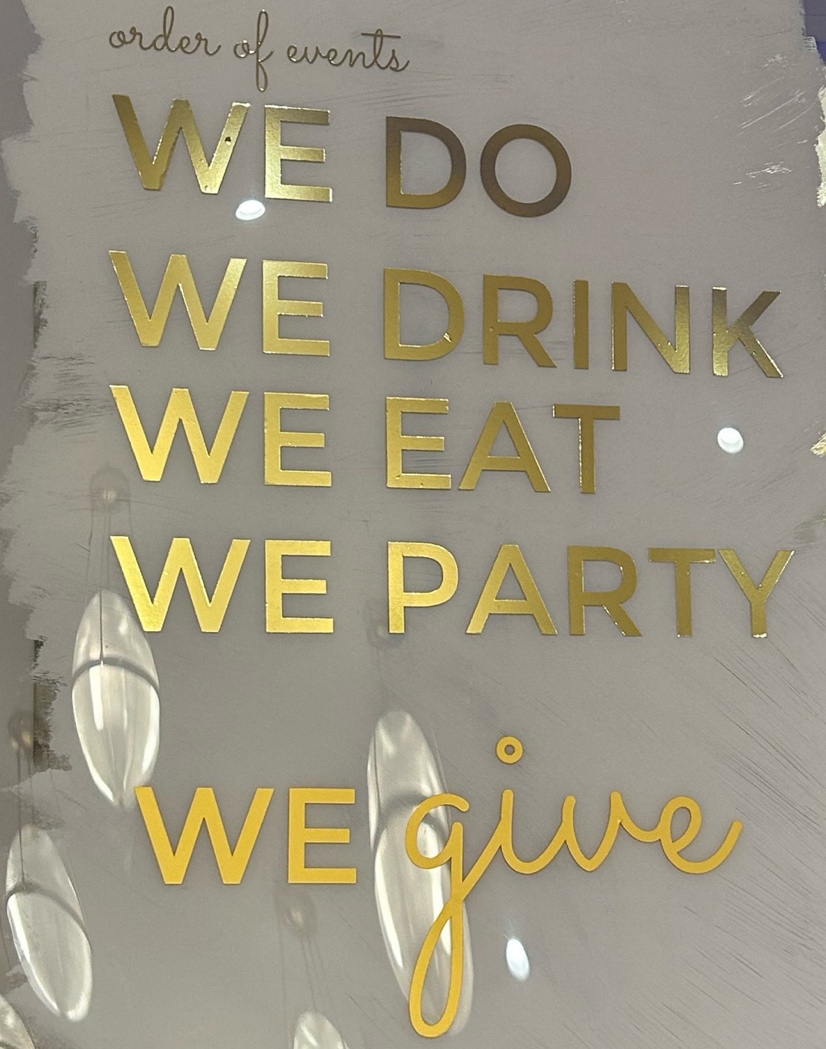 A sign with gold text on it saying we do, we drink, we eat, we party, we give is a great wedding theme fundraiser venue decor idea