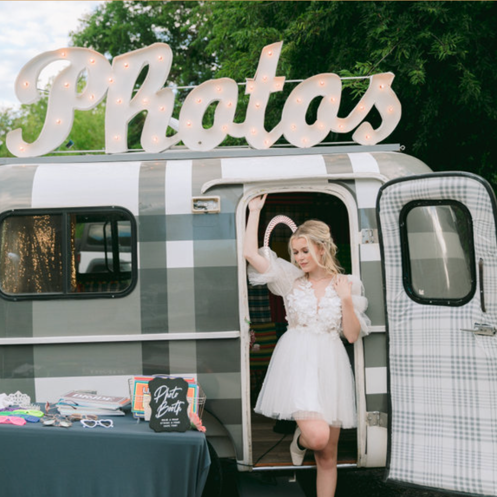 A person in a white dress standing in the door of a camper trailer for a photo tralier photo booth.